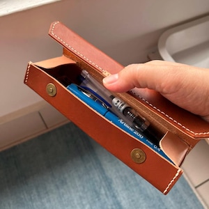 May include: A tan leather pen case with a magnetic clasp, containing a blue pen and a clear pen. The rectangular case features white stitching around the edges. The pen case is open, displaying the pens within.
