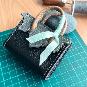 Puede incluir: Un pequeño bolso de mano de cuero negro con una cinta azul claro y un ribete de cuero gris. El bolso está sobre una estera de corte verde.