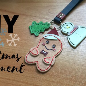 May include: DIY Christmas ornament with a gingerbread man, snowman, and a Christmas tree. The ornament is made of leather and has a red and white string attached to it. The text "Happy Holidays" is written on a green circle in the top right corner of the image.