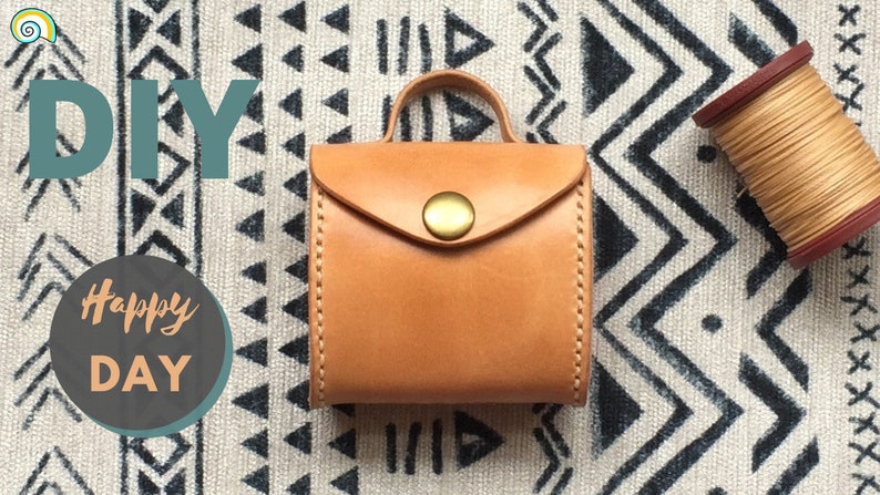 DIY project featuring a small, light brown leather storage bag with a flap closure and a brass button. The bag has a handle and is sitting on a white and black patterned fabric. A spool of beige thread is visible in the background.