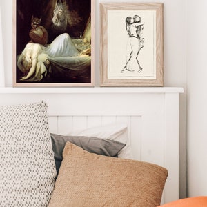 May include: Two framed art prints, one depicting a woman sleeping with a demon and a horse, the other depicting a couple embracing. The prints are hanging on a white wall above a bed with pillows.