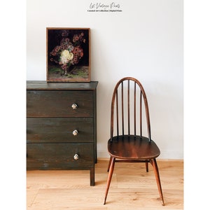 May include: A dark wood dresser with three drawers and a framed print of a floral still life above it. A dark wood chair with a curved back and spindle design sits to the right of the dresser. The chair and dresser are in front of a white wall.