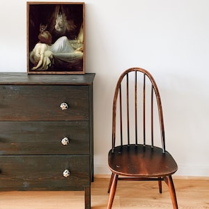May include: A vintage framed print of a woman sleeping in a bed with a demon figure standing over her. The print is hanging on a dark wooden dresser with three drawers. A wooden chair is in the foreground.