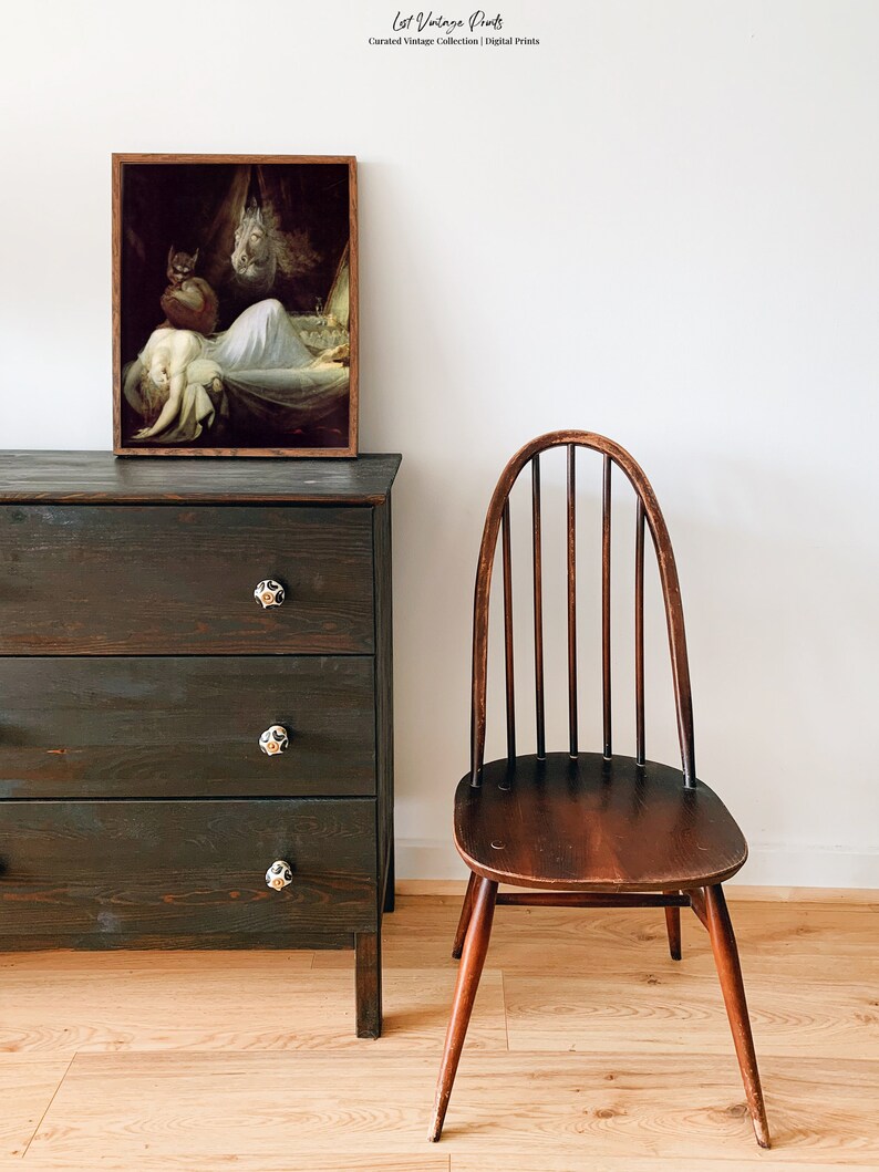 May include: A vintage framed print of a woman sleeping in a bed with a demon figure standing over her. The print is hanging on a dark wooden dresser with three drawers. A wooden chair is in the foreground.