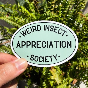 May include: An oval sticker with the text "WEIRD INSECT APPRECIATION SOCIETY" in black on a light green background. The sticker is held in front of a background of green plants.