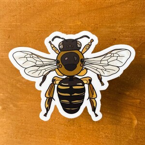 May include: A sticker featuring a stylized bee illustration. The bee has a brown and yellow body with black stripes, transparent wings, and a white border. The sticker is placed on a wooden surface.