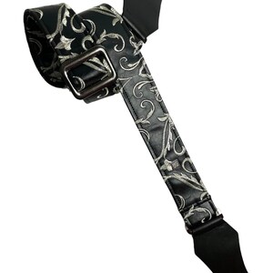 May include: A black guitar strap featuring a silver-toned embroidered floral scroll pattern throughout the length of the strap. It includes a rectangular metal adjustment buckle and solid black leather-like end tabs with a single hole for attachment.