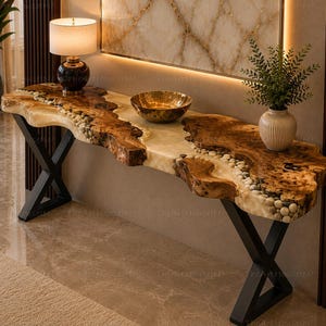 Peut inclure: Une table console dotée d'un plateau en bois à bord brut avec une rivière centrale en résine translucide incrustée de galets lisses. La table est soutenue par deux pieds en métal noir en forme de X et présente une finition en bois naturel.