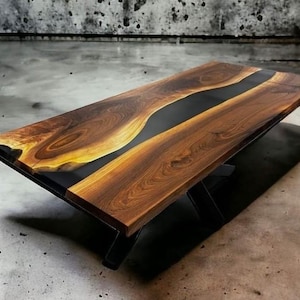 Op de afbeelding: Een lange, rechthoekige houten tafel met een zwarte epoxyrivier die door het midden loopt. De tafel wordt ondersteund door een zwarte metalen basis.