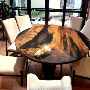 Pode incluir: Uma mesa de jantar redonda de madeira com uma incrustação de resina epóxi preta. A mesa tem um acabamento de madeira natural e é cercada por cadeiras brancas.