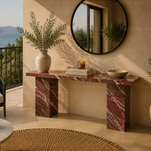 May include: A rectangular console table made of deep red marble with white veining, featuring two thick slab legs. The table surface holds a beige ceramic vase with greenery, two stacked books, a glass candle, and a shallow beige bowl.