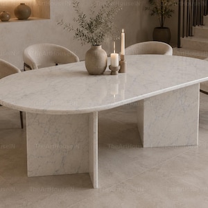 May include: An oval dining table with a white marble top featuring grey veining, supported by two thick, rectangular marble pedestal bases. Four curved, cream-coloured upholstered chairs with dark wooden legs are positioned around the table.