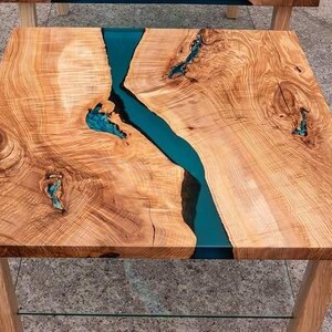 May include: Square wooden side table with a natural wood grain finish and a vibrant teal resin river running through the center. The table has light-colored wooden legs and a clear glass shelf.