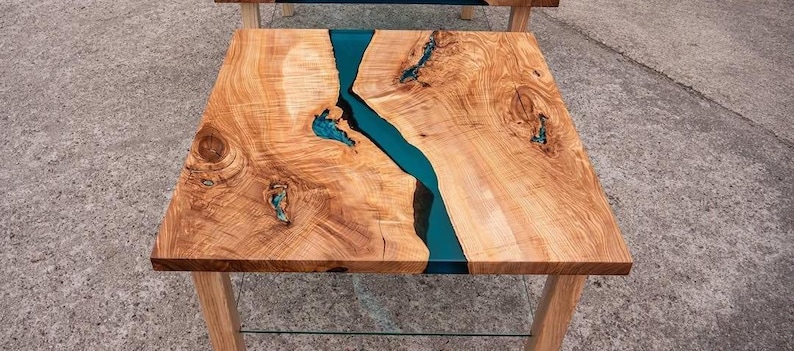 May include: Square wooden side table with a natural wood grain finish and a vibrant teal resin river running through the center. The table has light-colored wooden legs and a clear glass shelf.