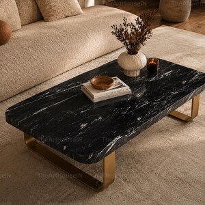 Puede incluir: Una mesa de centro rectangular con una superficie de mármol negro con vetas blancas y bordes redondeados. La mesa se apoya sobre dos patas metálicas curvas de tono dorado que proporcionan una base estable para la pesada superficie de piedra.