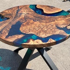 May include: Circular wooden table with a distinctive design. The tabletop has a natural wood grain pattern with blue and teal resin accents. The table is supported by a black metal base. A modern piece of furniture.