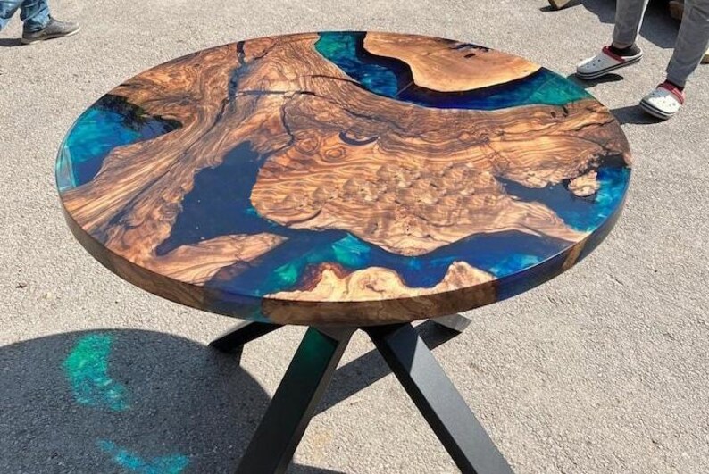 May include: Circular wooden table with a distinctive design. The tabletop has a natural wood grain pattern with blue and teal resin accents. The table is supported by a black metal base. A modern piece of furniture.