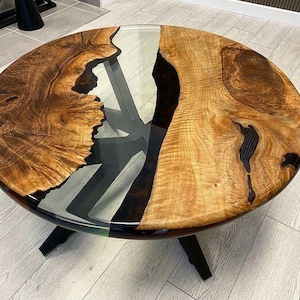 Puede incluir: Una mesa de centro redonda de madera con un río de resina epoxi negra que fluye por el centro. La mesa tiene una tapa de vidrio transparente y una base de metal negro.