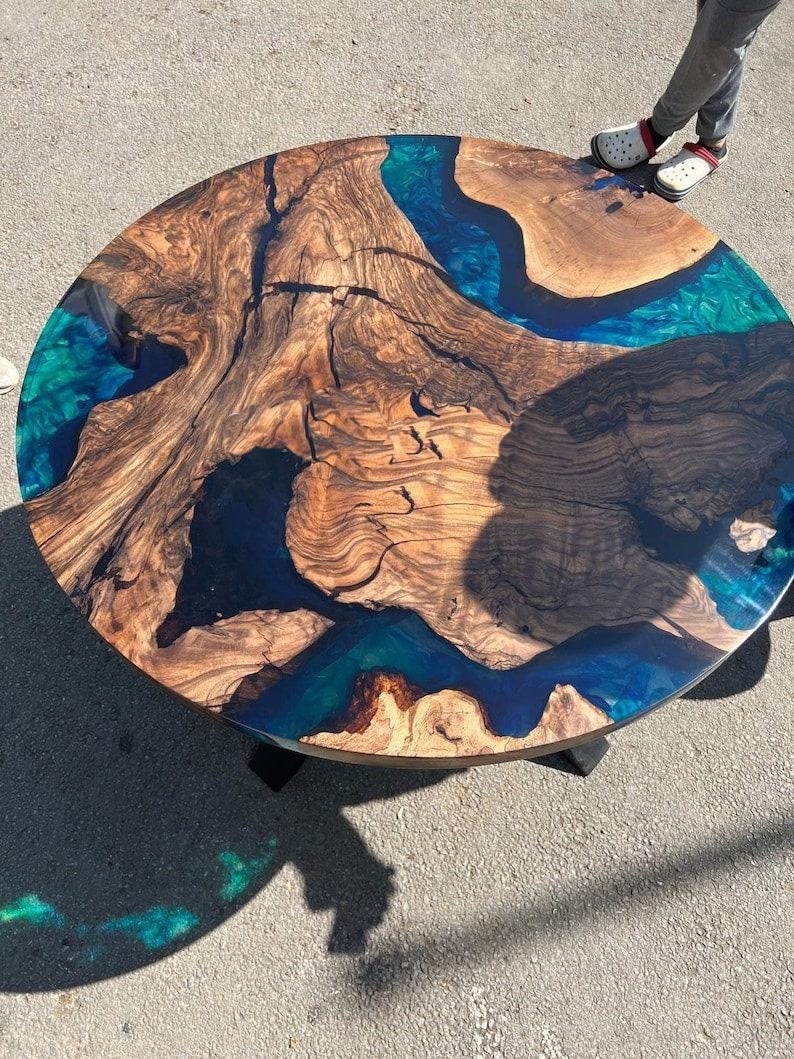 May include: Circular coffee table crafted from wood, featuring a river design made with blue and teal epoxy resin. The table showcases a natural wood grain pattern, complemented by the vibrant resin. It stands on a black base.