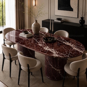 May include: An oval dining table with a deep red marble top featuring white veining and a fluted cylindrical base, surrounded by six cream-colored upholstered chairs with dark wooden legs, arranged on a textured beige area rug.