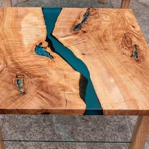 May include: Square wooden coffee table with a teal epoxy resin river running through the center. The table has a natural wood grain finish and sits on four wooden legs. The table is approximately 30 inches square.