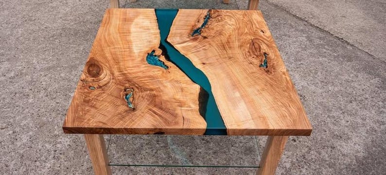 May include: Square wooden coffee table with a teal epoxy resin river running through the center. The table has a natural wood grain finish and sits on four wooden legs. The table is approximately 30 inches square.