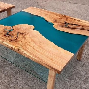 May include: A handcrafted wooden coffee table with a teal epoxy resin river. The table features natural wood grain patterns and a clear glass shelf. The table's dimensions are approximately 48 inches long, 24 inches wide, and 18 inches high.