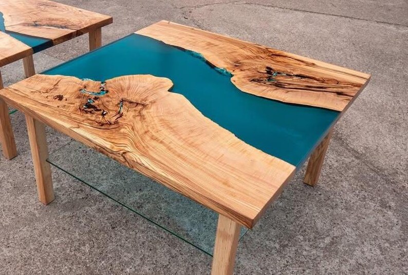 May include: A handcrafted wooden coffee table with a teal epoxy resin river. The table features natural wood grain patterns and a clear glass shelf. The table's dimensions are approximately 48 inches long, 24 inches wide, and 18 inches high.