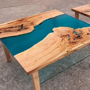May include: Two wooden coffee tables with a teal epoxy resin river design. The tables have natural wood grain and are supported by light-colored wooden legs. One table has a glass shelf.
