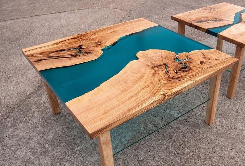 May include: Two wooden coffee tables with a teal epoxy resin river design. The tables have natural wood grain and are supported by light-colored wooden legs. One table has a glass shelf.
