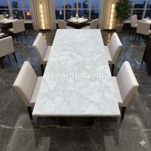 Puede incluir: Una mesa de comedor rectangular con una superficie blanca similar al mármol, rodeada de seis sillas color crema. La mesa es el punto focal, con el texto "TheArtHouseIN" visible. El entorno es un restaurante moderno.
