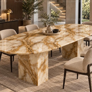 Puede incluir: Una mesa de comedor ovalada con una superficie de mármol pulido en tonos blanco y ámbar con vetas prominentes, sostenida por dos pedestales rectangulares de mármol a juego. La mesa está rodeada por cuatro sillas con tapizado texturizado color crema.