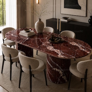 May include: An oval dining table with a deep red marble top and a matching fluted cylindrical base, surrounded by six beige upholstered chairs with dark wooden legs. The table surface features white veining and holds a vase and candles.