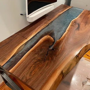 May include: A handcrafted wooden table with a live edge design. The table features a river of blue resin flowing through the centre, creating a unique visual effect. The wood has a rich, dark brown colour, and the table is supported by a metal frame.