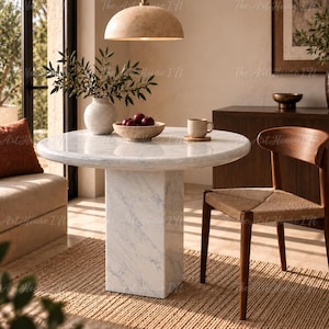 Puede incluir: Una mesa de comedor redonda de mármol blanco con una base de pedestal gruesa, acompañada de una silla de madera con asiento tejido. Sobre la mesa hay un jarrón de cerámica con ramas verdes, un cuenco con frutas rojas y una taza pequeña.