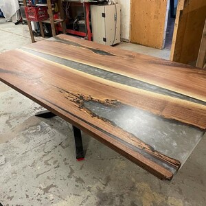 May include: A large, rectangular wooden table with a clear epoxy resin river running down the center. The table has a dark brown finish and sits on a black metal base. The table's dimensions are approximately 72 inches long and 36 inches wide.