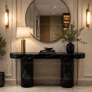 Può includere: Un tavolo console in marmo nero con due basi cilindriche scanalate, sormontato da una lampada dorata, una pila di libri con l'etichetta Chanel, una ciotola dorata e un vaso nero con fogliame verde, posizionato sotto un grande specchio tondo.