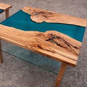 May include: A wooden coffee table with a teal epoxy resin river design. The table has natural wood grain and is supported by four wooden legs. The table measures approximately 48 inches long and 24 inches wide.