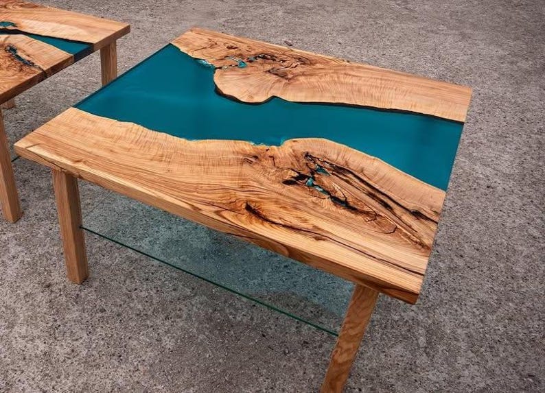 May include: A wooden coffee table with a teal epoxy resin river design. The table has natural wood grain and is supported by four wooden legs. The table measures approximately 48 inches long and 24 inches wide.