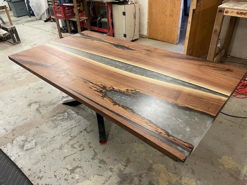 May include: A large, rectangular wooden table with a clear epoxy resin river running down the center. The table has a dark brown finish and sits on black metal legs. The table's dimensions are approximately 72 inches long and 36 inches wide.