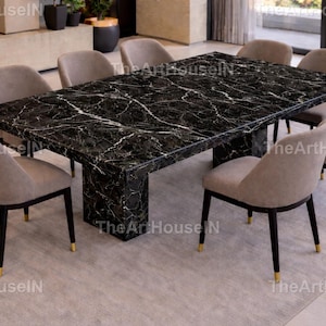 Puede incluir: Una mesa de comedor moderna con tapa de mármol negro y base rectangular. Ocho sillas tapizadas beige con patas oscuras y detalles dorados rodean la mesa. El texto The ArtHouseIN es visible.