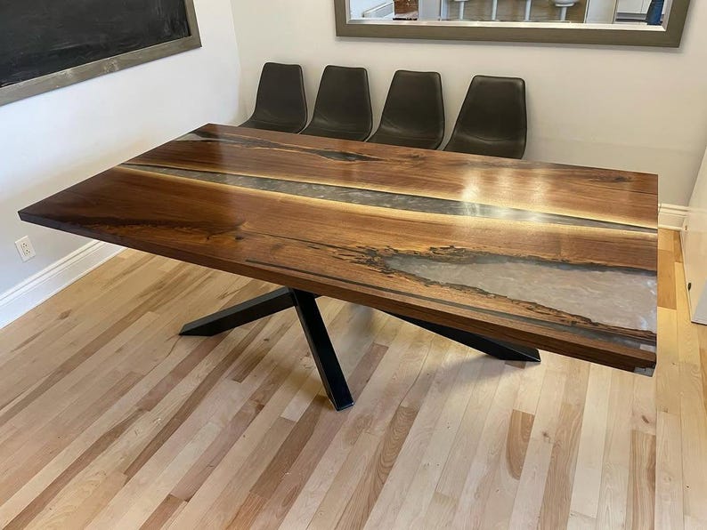 May include: A large, rectangular wooden dining table with a dark brown finish and a clear, light-colored epoxy river running down the center. The table has a black metal base and is set in a room with hardwood floors.