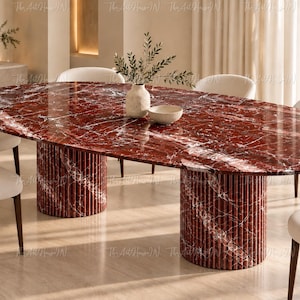 Op de afbeelding: Een ovale eettafel met een gepolijst dieprood marmeren blad met witte aders, ondersteund door twee cilindrische bases met een verticale gegroefde textuur. De tafel is omringd door zes gebroken witte gestoffeerde stoelen met donkere houten poten.