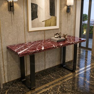 May include: A rectangular console table featuring a polished deep red marble top with white veining, supported by two black U-shaped metal legs. The table holds a stack of two books and a small metallic decorative object on its surface.