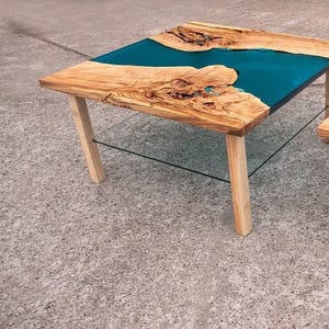 May include: A handcrafted wooden coffee table with a teal epoxy resin river design. The table features a natural wood grain top and four light-colored wooden legs. A clear glass shelf is positioned below the tabletop.