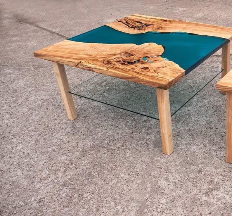May include: A handcrafted wooden coffee table with a teal epoxy resin river design. The table features a natural wood grain top and four light-colored wooden legs. A clear glass shelf is positioned below the tabletop.