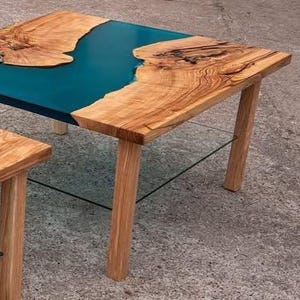 May include: Two wooden tables with a teal epoxy resin river design. The tables have natural wood edges and light brown wooden legs. The larger table is in the background, and a smaller table is in the foreground.