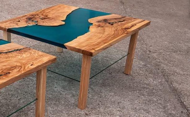 May include: Two wooden tables with a teal epoxy resin river design. The tables have natural wood edges and light brown wooden legs. The larger table is in the background, and a smaller table is in the foreground.