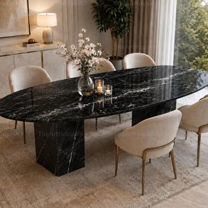 Puede incluir: Una mesa de comedor ovalada con tablero de mármol negro y base de pedestal a juego, rodeada de seis sillas tapizadas en color crema con patas de metal dorado. Sobre la mesa hay un jarrón de cristal con flores blancas y dos portavelas.