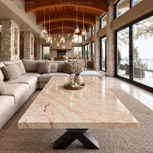 Puede incluir: Una mesa de centro rectangular con una superficie de mármol beige pulido con vetas marrones y una base de metal negro en forma de X. La mesa se asienta sobre una alfombra marrón texturizada en una habitación con un sofá seccional, pilares de piedra y lámparas colgantes de cristal.